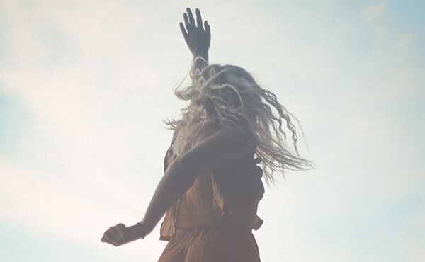 Woman with flowing hair raising her arms to the sky, symbolising positive energy and freedom against a soft blue background.
